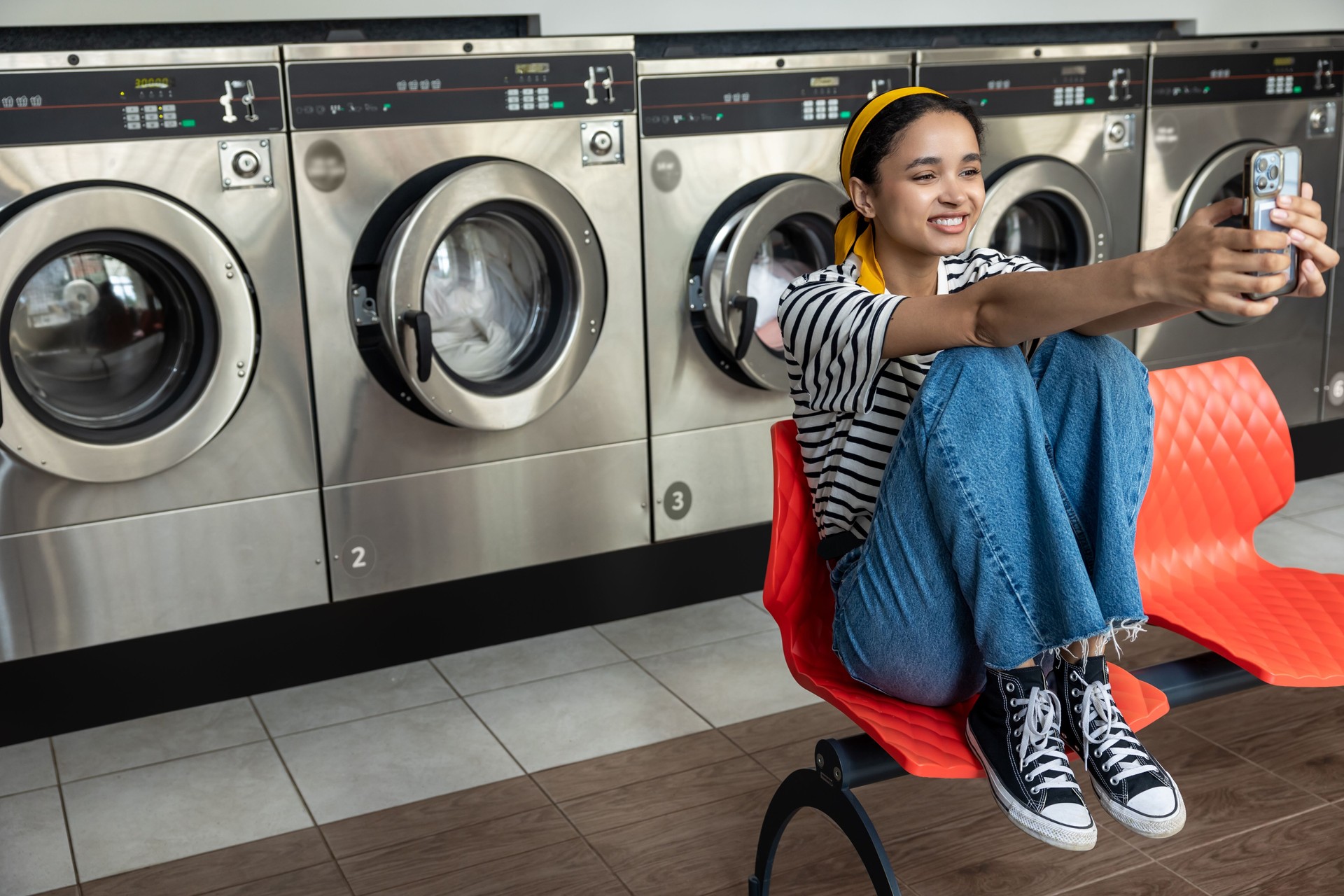 Housewife in laundromat taking selfie on mobile phone while using washing machine